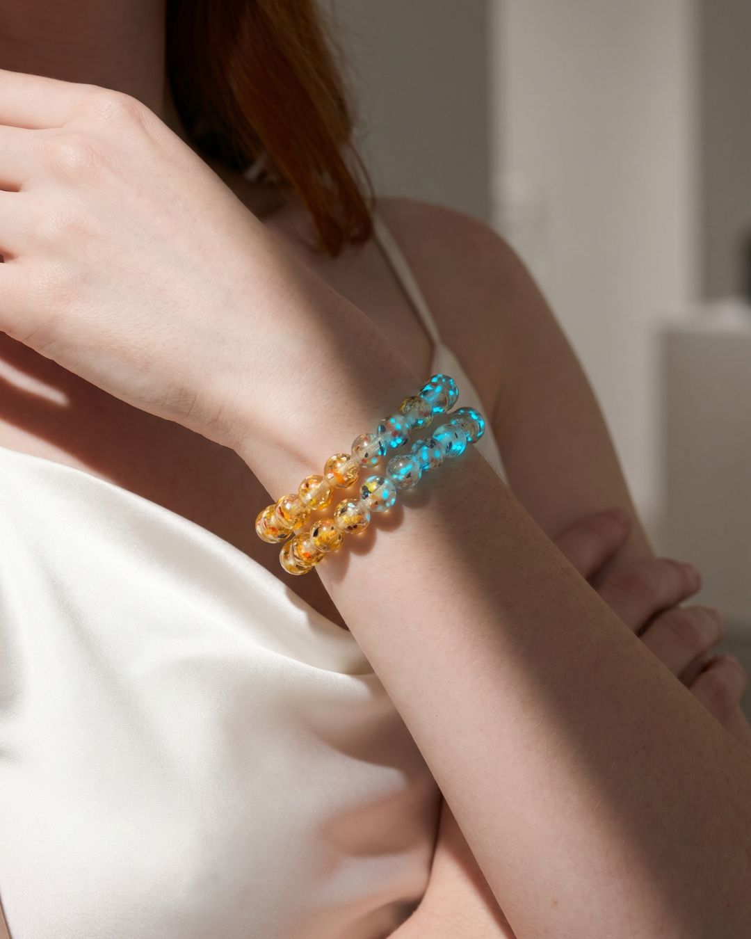 Stacked translucent glass bead bracelets in turquoise blue and amber yellow, styled on a wrist with a white satin top.