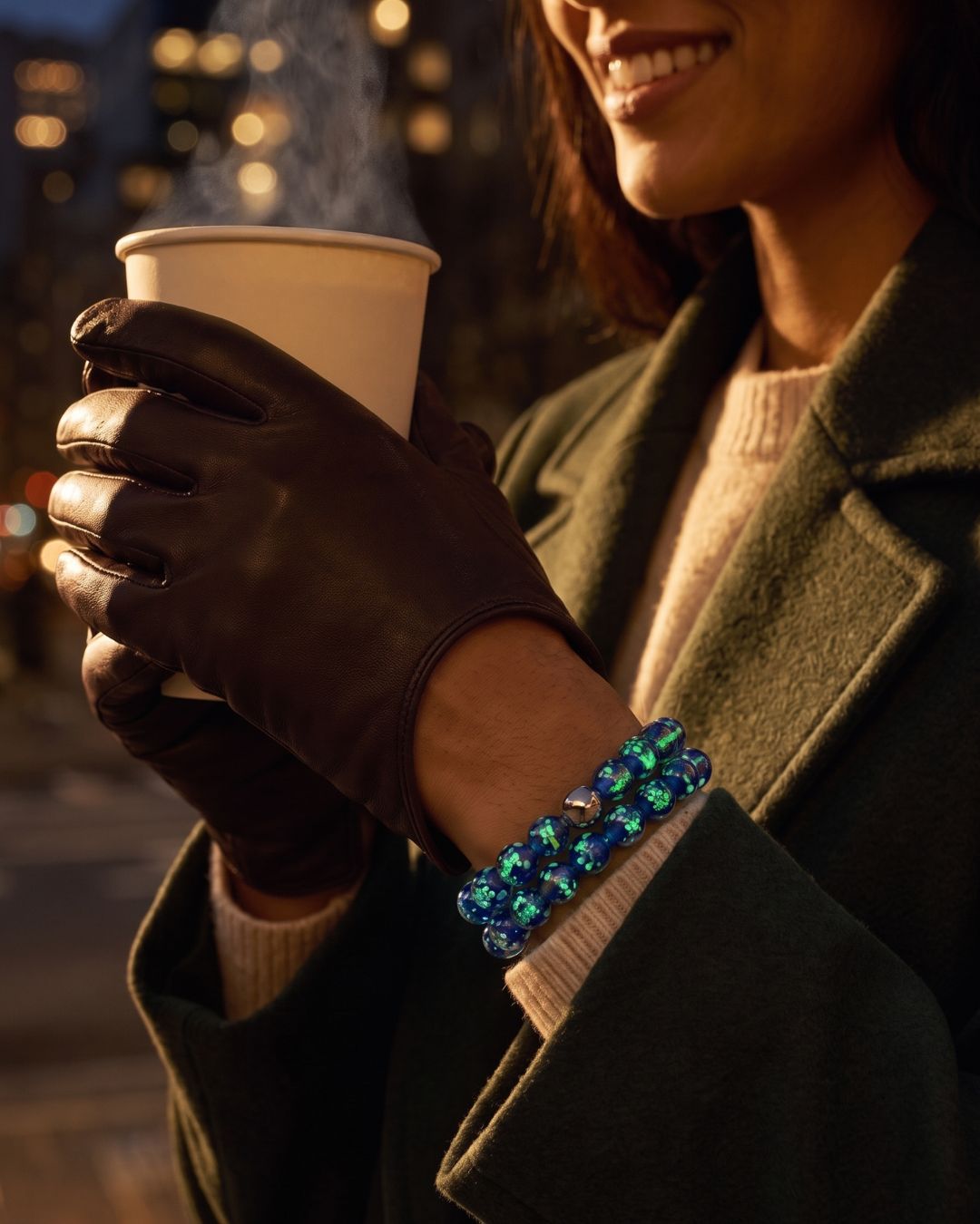 NOGU blue and green iridescent beaded bracelet styled with brown leather gloves and a dark green coat.