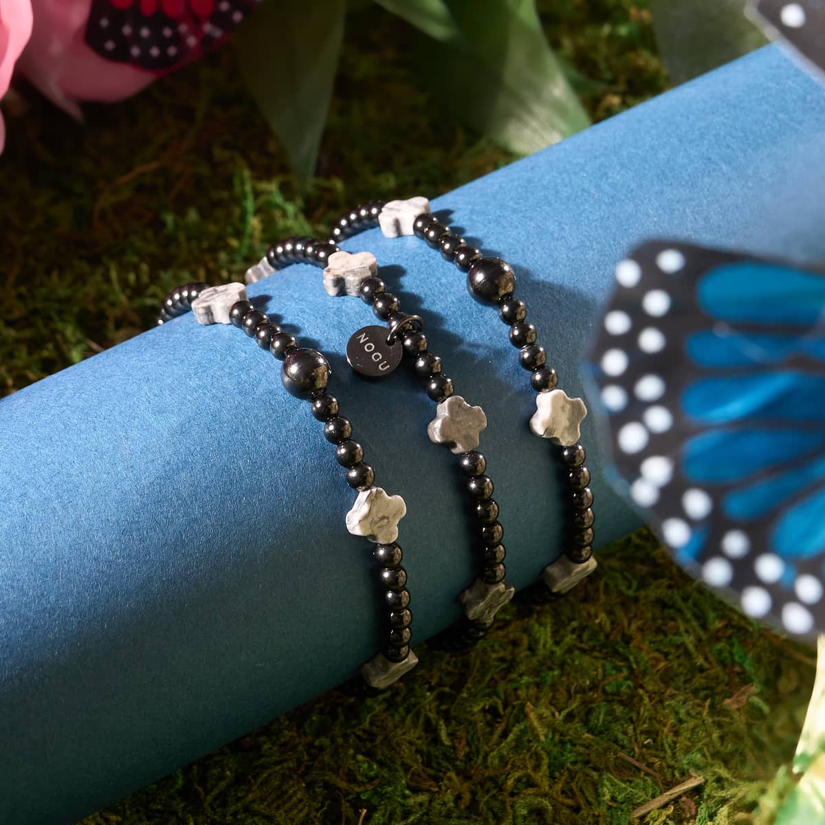 Luck in the Dark Gunmetal Clover Essence Bracelet set with black beads grey clover charms and NOGU tag on blue cylinder background
