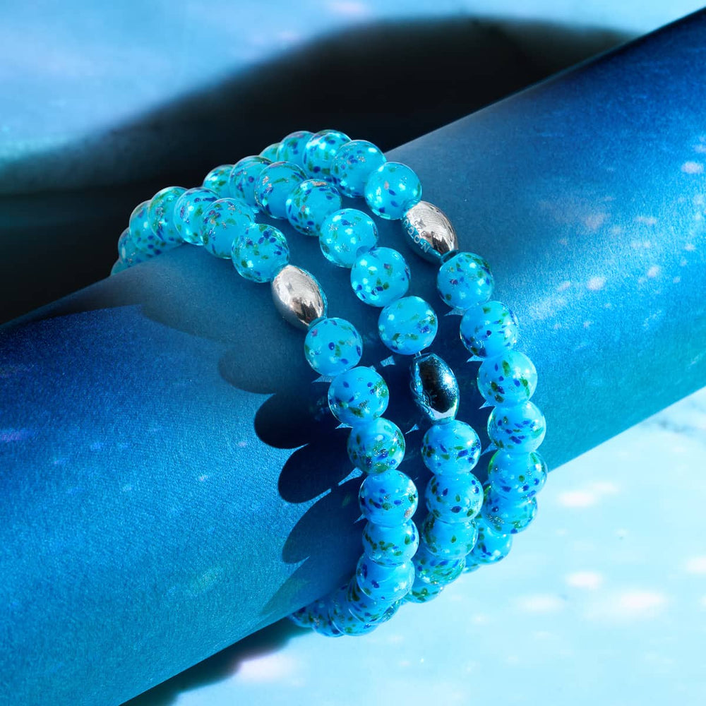 Blue Juniper Firefly Glass Bracelet with blue speckled beads and sterling silver clasp coiled on blue surface by NOGU